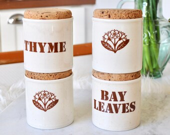 Op de afbeelding: Vier witte keramische kruidenpotten met kurken deksels. De potten zijn gelabeld met "THYME", "BAY LEAVES" en hebben bloemmotieven.