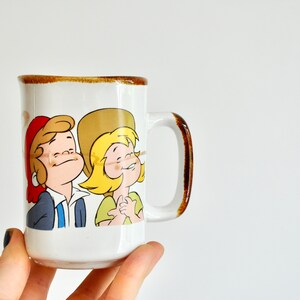 K&ouml;nnte beinhalten: Wei&szlig;e Keramiktasse mit braunem Rand und Henkel. Die Tasse zeigt eine Comic-Illustration von zwei Kindern mit blonden Haaren, eines tr&auml;gt einen roten Hut und das andere einen braunen Hut.