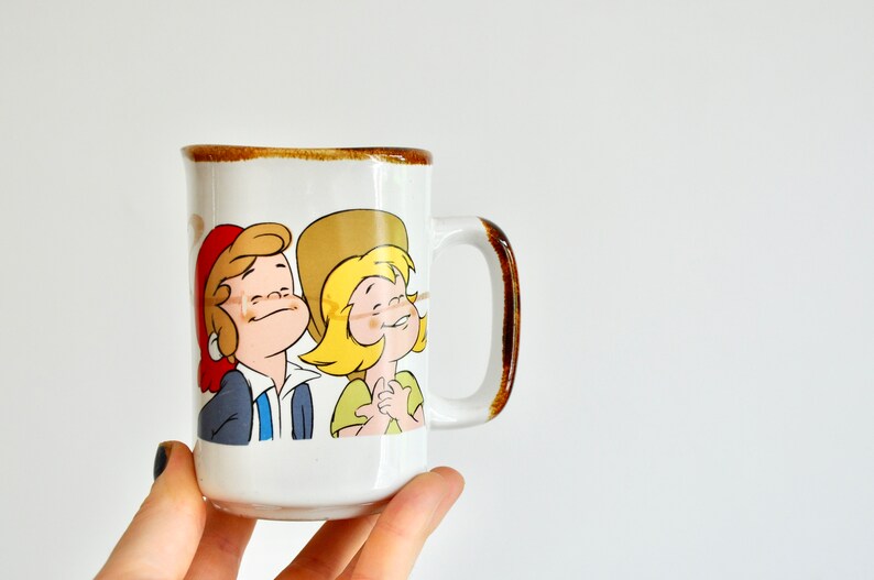 K&ouml;nnte beinhalten: Wei&szlig;e Keramiktasse mit braunem Rand und Henkel. Die Tasse zeigt eine Comic-Illustration von zwei Kindern mit blonden Haaren, eines tr&auml;gt einen roten Hut und das andere einen braunen Hut.