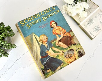 May include: A vintage book titled "Schoolgirls Story Bumper" with a colorful illustration of two girls camping and cooking over a fire. The book cover features the text "The Evening Meal" in a yellow banner.