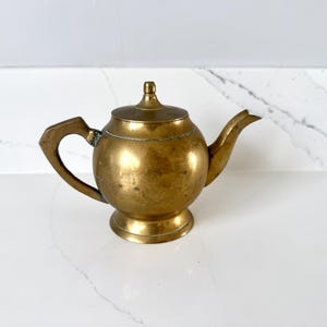 Puede incluir: Una tetera vintage de latón con cuerpo redondeado, boquilla curva y asa geométrica. La tapa está rematada con un pequeño pomo. La tetera tiene un color dorado cálido y se asienta sobre una pequeña base.