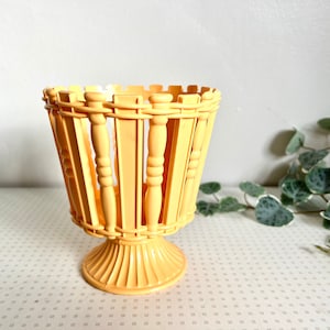 May include: A light yellow, pedestal basket with a woven design. The basket has a flared top and a decorative base. The vertical slats and rounded supports give it a vintage look. The basket is likely made of plastic or a similar material.