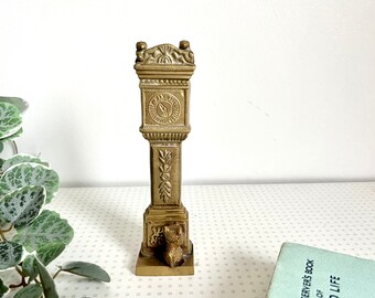 May include: A gold-toned brass clock tower figurine with a small dog figurine at the base. The clock face has Roman numerals and a black hand.