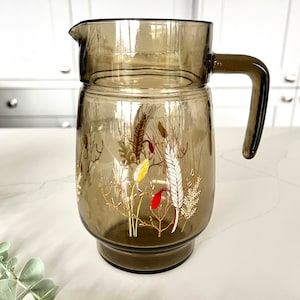 May include: A brown glass jug with a floral design featuring wheat stalks and red berries. The jug has a wide mouth and a handle.