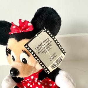以下が含まれることがあります： 赤と白のドット柄のドレスと赤いリボンをつけた、ミニーマウスのぬいぐるみです。おもちゃには、黒と白のタグが付いており、「注意：このタグを完全に取り外し、おもちゃを子供に渡す前に留め具を捨ててください。 0122-19459 &copy;DISNEY」というテキストが書かれています。