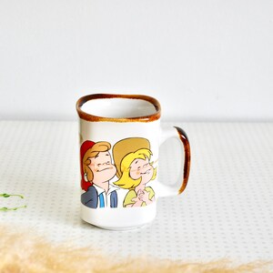 K&ouml;nnte beinhalten: Wei&szlig;e Keramiktasse mit braunem Rand und Henkel. Die Tasse zeigt eine Comic-Illustration eines Mannes und einer Frau in Rot, Gelb und Blau.