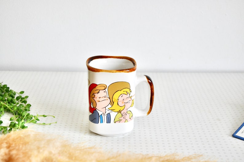 K&ouml;nnte beinhalten: Wei&szlig;e Keramiktasse mit braunem Rand und Henkel. Die Tasse zeigt eine Comic-Illustration eines Mannes und einer Frau in Rot, Gelb und Blau.