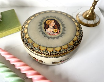 May include: A round, cream-colored trinket box with a gold and brown decorative border. The lid features a portrait of a woman with a yellow rose in a gold oval frame.