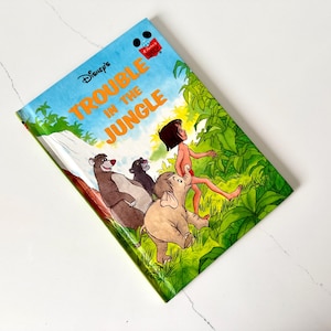 May include: A vintage Disney's "Trouble in the Jungle" book. The cover features illustrations of Baloo the bear, Bagheera the panther, an elephant, and Mowgli. The title is in orange and yellow letters against a blue sky background.