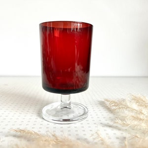 May include: A dark red stemmed glass with a clear glass base. The glass is sitting on a white surface with a white fluffy plant in the background.