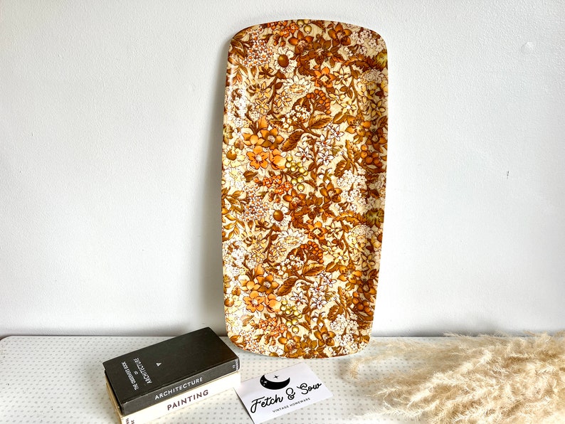 May include: A vintage serving tray with a floral pattern in shades of brown, orange, and white. The tray is made of a hard plastic material and has a rectangular shape.