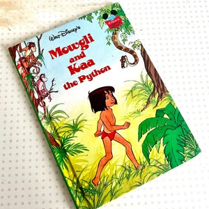 Op de afbeelding: Een vintage kinderboekomslag met een cartoonillustratie van Mowgli uit Disney's Jungle Book. De omslag is groen en heeft de titel "Mowgli en Kaa de Python".