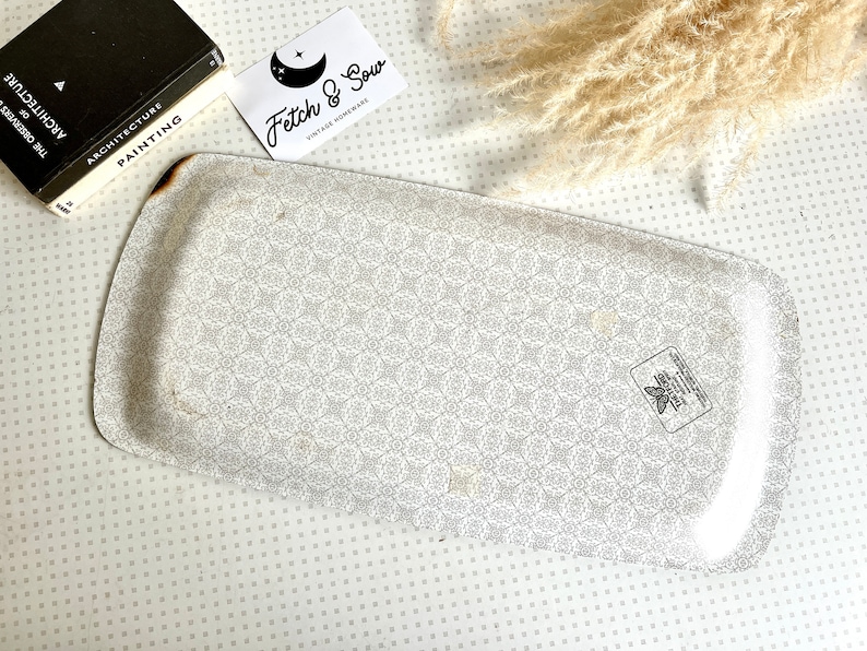 May include: A white metal serving tray with a delicate floral pattern. The tray has a slightly rounded shape and a raised edge. The bottom of the tray has a small black and white sticker with a butterfly design.