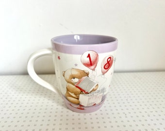 May include: A purple and white ceramic mug with a cartoon bear holding a gift box. The mug has a handle and features red balloons with the numbers "18" printed on them.