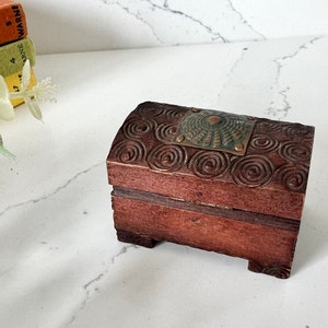 Può includere: Un piccolo forziere in legno con finitura marrone scuro. Il coperchio è adornato con disegni a spirale intagliati e una placca metallica decorativa. Il forziere è rettangolare con piedini e la placca metallica ha un motivo a raggiera.