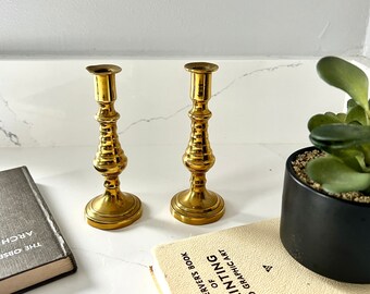 May include: Two gold metal candle holders with a textured design. The candle holders are on a white surface with a book and a potted plant.