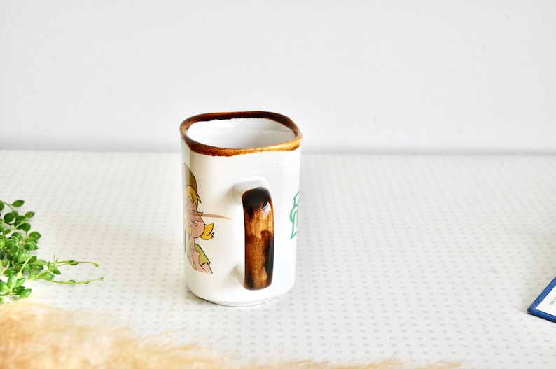 K&ouml;nnte beinhalten: Wei&szlig;e Keramiktasse mit braunem Henkel und einer Comic-Illustration eines M&auml;dchens mit blonden Haaren. Die Tasse hat einen braunen Rand und eine gr&uuml;ne Aufschrift.