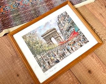 May include: A framed print of a Parisian street scene featuring the Arc de Triomphe. The artwork is in a light brown wooden frame and depicts a bustling street with people, shops, and trees. The print has a white border.
