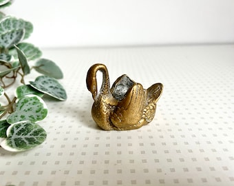 May include: A small, antique brass swan figurine. The swan is in a seated position with its neck curved gracefully. The brass has a warm, aged patina. The swan's wings and feathers are detailed. A decorative item.