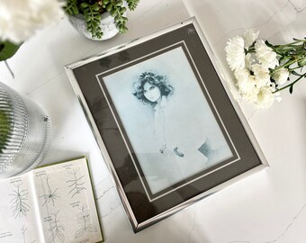 May include: A framed black and white photograph of a woman with curly hair. The frame is silver with a brown mat.