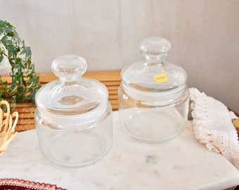 May include: Two clear glass jars with lids on a white marble surface. The jars are empty and have a slightly frosted look.