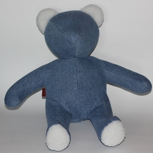 Puede incluir: Un oso de peluche de mezclilla azul con orejas y pies de piel blanca. El oso está sentado sobre una superficie blanca.