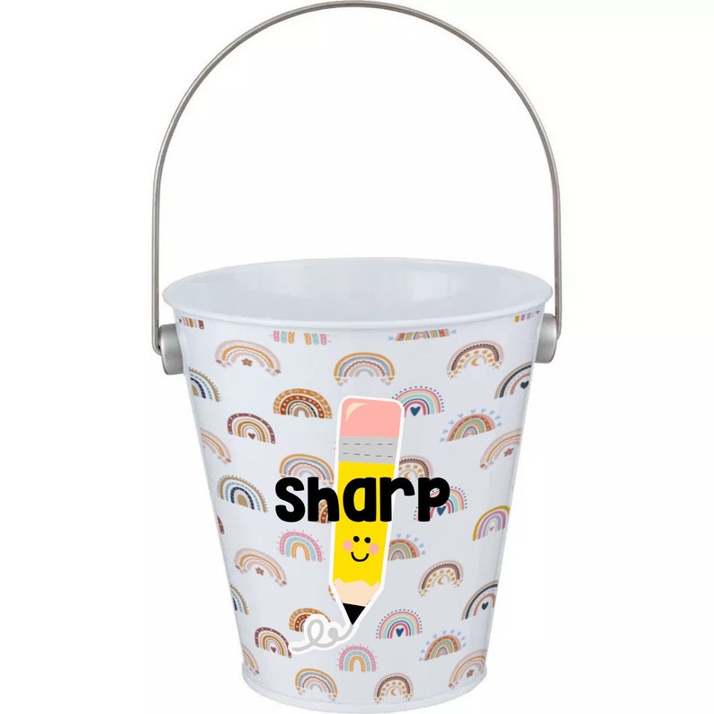 Puede incluir: Un cubo de metal blanco con un asa plateada. El cubo est&aacute; decorado con un patr&oacute;n de arco&iacute;ris en varios colores. Un l&aacute;piz amarillo con una goma de borrar rosa y una cara sonriente est&aacute; en la parte delantera del cubo. La palabra "sharp" est&aacute; escrita en letras negras debajo del l&aacute;piz.