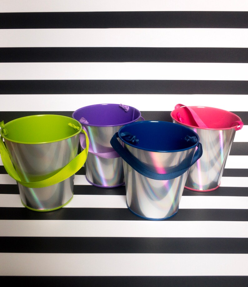 Puede incluir: Cuatro cubos peque&ntilde;os de plata con acabados iridiscentes y asas de colores. Los cubos est&aacute;n dispuestos en fila sobre un fondo de rayas blancas y negras. Las asas son verdes, moradas, azules y rosas.