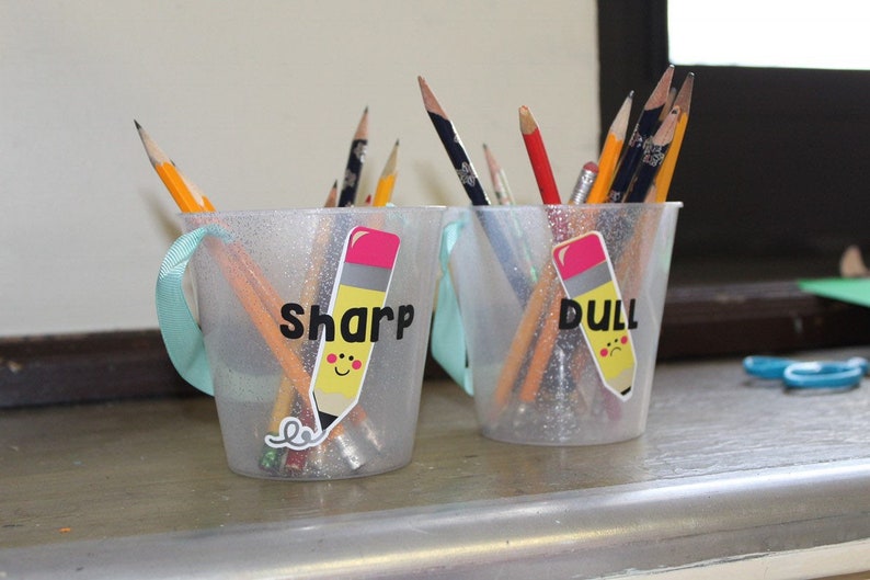 Puede incluir: Dos vasos de pl&aacute;stico transparente con asas, llenos de l&aacute;pices. Un vaso est&aacute; etiquetado como "Sharp" y el otro como "Dull".