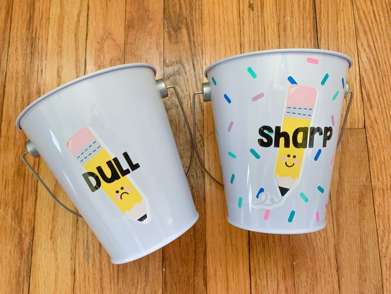 Puede incluir: Dos cubos met&aacute;licos peque&ntilde;os con asas. Un cubo est&aacute; etiquetado como "DULL" con un l&aacute;piz amarillo con cara triste. El otro cubo est&aacute; etiquetado como "SHARP" con un l&aacute;piz amarillo con cara feliz. Ambos cubos tienen un patr&oacute;n de confeti.