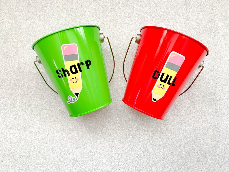 Puede incluir: Dos cubos peque&ntilde;os de metal verde y rojo con dibujos de l&aacute;pices en ellos. El cubo verde dice "sharp" y el cubo rojo dice "dull".