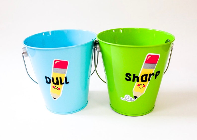 Puede incluir: Dos cubos met&aacute;licos peque&ntilde;os, uno azul y otro verde, con pegatinas en forma de l&aacute;piz. El cubo azul dice "DULL" y el cubo verde dice "SHARP".