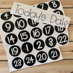 May include: A set of black and white circular stickers with numbers 1 through 24. The stickers are arranged in a grid pattern. A white banner with the text "Today's Date" is placed on top of the stickers.