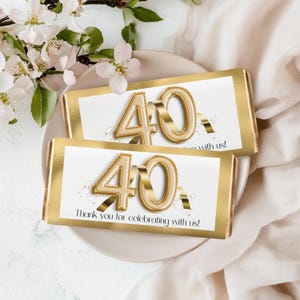 Könnte beinhalten: Zwei goldfarbene Schokoriegel mit einem weißen Etikett mit einem goldenen 40-Ballon-Design und dem Text "Vielen Dank, dass Sie mit uns feiern!". Die Riegel liegen auf einem weißen Teller mit Blumen.
