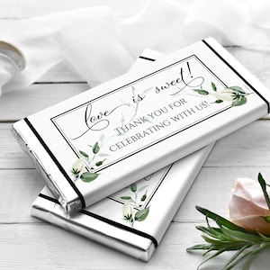 May include: Two white chocolate bars with silver foil wrappers. The top bar has a black border around a white rectangle with the text "Love is sweet! Thank you for celebrating with us!" in black cursive font. The bottom bar has a black border around a white rectangle with a floral design in white and green.