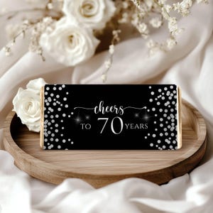 Op de afbeelding: Een chocoladereep met een zwarte wikkel met de tekst "Cheers to 70 Years" in witte letters. Zilveren confetti stippen omlijsten de tekst. De reep ligt op een houten dienblad, met witte bloemen op de achtergrond.