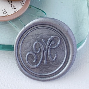 May include: A silver wax seal with the letter "M" in a cursive font. The seal is on a clear glass surface with a dragonfly design. There are three round adhesive labels with instructions for removing the liner to expose the adhesive.