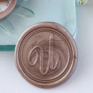 May include: A close-up of a gold wax seal with the letter "U" in a cursive script. The wax seal is on a light blue glass surface.