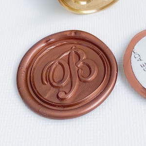 May include: A gold-colored wax seal stamp with a wooden handle and a copper-colored wax seal. The stamp has the letter "B" in an elegant script. A round sticker with text is also visible.