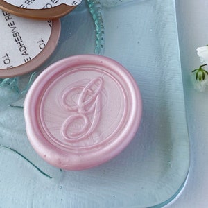 May include: A pink wax seal with a letter G in the center, on a light blue glass surface with a dragonfly design. There are three round adhesive labels with instructions to remove the liner to expose the adhesive.