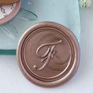 May include: A rose gold wax seal with the letter "F" in a cursive font. The seal is on a clear glass surface with a dragonfly design. There are three adhesive liners with instructions to remove the liner to expose the adhesive.
