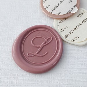 May include: A pink wax seal with the letter L in a cursive font. The seal is on a white surface with two white adhesive labels with text that reads "ADHESIVE REMOVE TO EXPOSE ADHESIVE REMOVE TO EXPOSE".