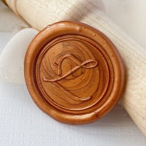 May include: A wooden handle with a gold band and a copper wax seal with a cursive letter L. The wax seal is on a white background.