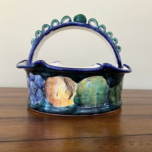 May include: A ceramic fruit basket with a blue and green glaze. The basket features a handle and is decorated with painted fruit, including grapes, peaches, and lemons.