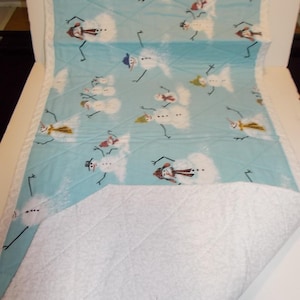 May include: A quilted, light blue blanket featuring a repeating pattern of snowmen with various hats and scarves. The snowmen are set against a background of white clouds. The blanket has a white, quilted backing and a white border.