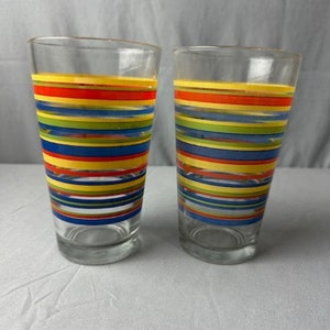 May include: Two clear glass drinking glasses with colorful striped designs. The stripes are yellow, red, blue, green, and orange.