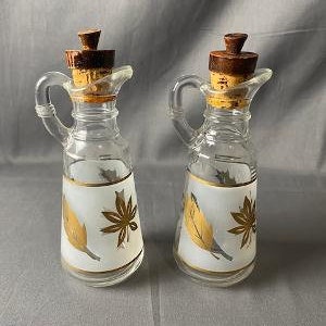 May include: Two clear glass cruets with handles and cork stoppers. Each bottle features a frosted white band around the center decorated with gold-colored leaf patterns and thin gold horizontal lines at the top and bottom of the frosted section.
