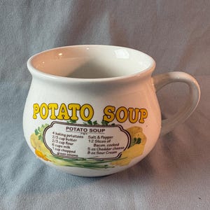 May include: A white ceramic soup mug with a handle, featuring the words "POTATO SOUP" in yellow. The mug includes a recipe for potato soup with ingredients like potatoes, butter, flour, milk, and cheese.