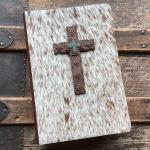 May include: A rectangular journal with a white and brown cowhide cover featuring a centered brown cross ornament. The cross has a dark brown textured finish with small diamond-shaped accents and a central turquoise and red cross-shaped inlay.