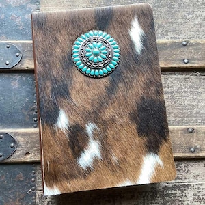 May include: A rectangular journal with a brown, black, and white cowhide cover. Centered on the front cover is a circular silver-toned concho ornament featuring a radial pattern of small, oval-shaped turquoise-colored stones set in metal.